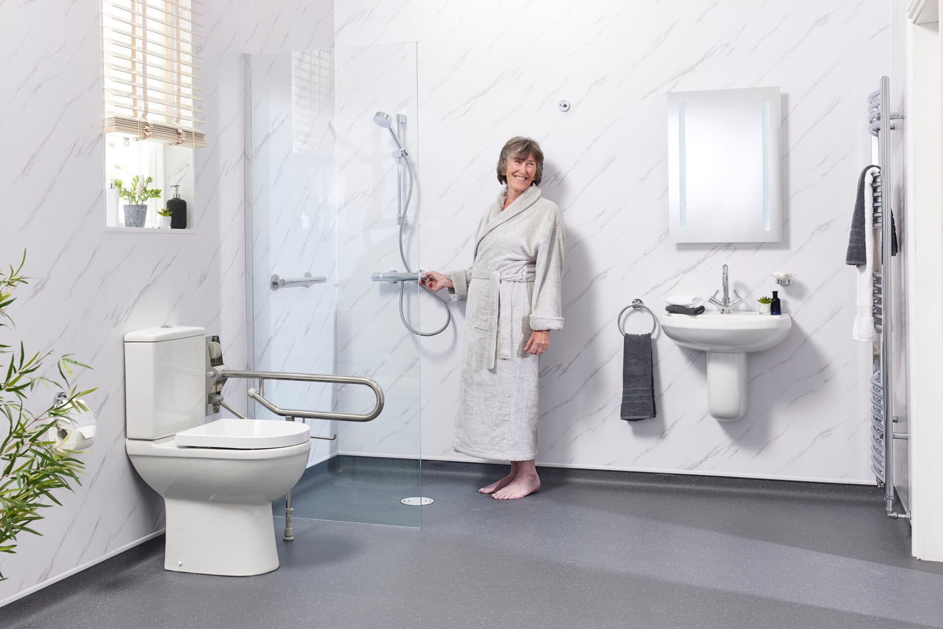An older woman standing at the controls of a level-access shower, with a grab rail beside the toilet and a level, non-slip floor.